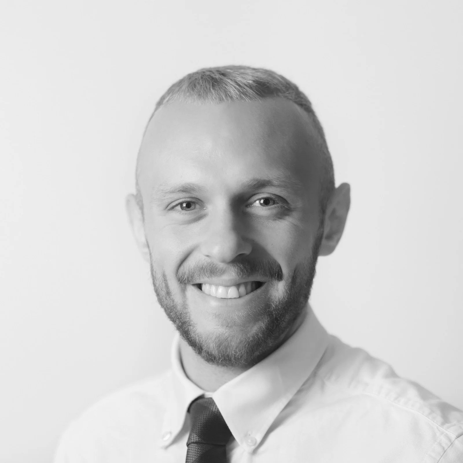 Black and white portrait of smiling man with beard, short hair, wearing a collared shirt and a tie.