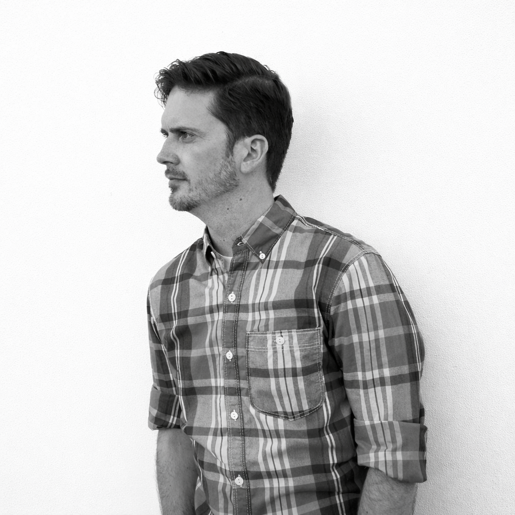 Black and white photo of a man with styled hair and a short beard, wearing a plaid button-up shirt, standing sideways against a plain white wall.